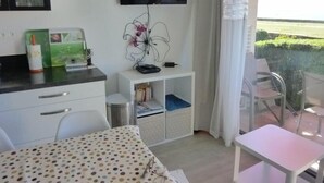 Ett modernt rum med ett matbord, en tv som är monterad på väggen, ett litet vitt skåp och en balkong med utsikt över grönskan.