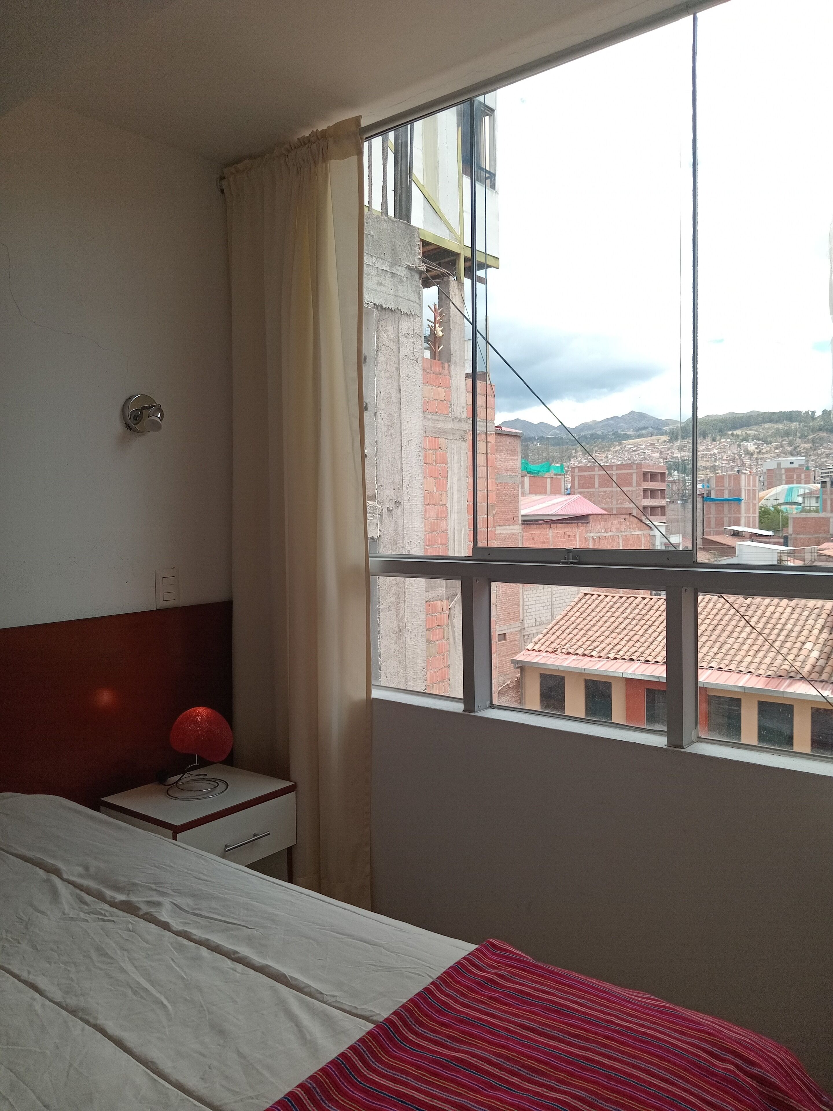 Ein Schlafzimmer mit Bett, Nachttisch mit Lampe und Fenster mit Blick auf Gebäude und Berge.