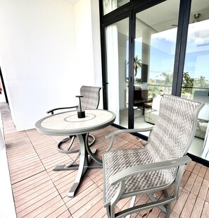 A balcony with a round table and two wicker chairs, a sliding glass door, and a view of palm trees and buildings.