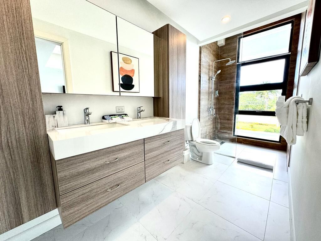 A modern bathroom with a large mirror, wooden cabinetry, a dual sink vanity, a shower, and a view of the outdoors.