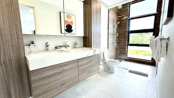 A modern bathroom with a large mirror, wooden cabinetry, a dual sink vanity, a shower, and a view of the outdoors.