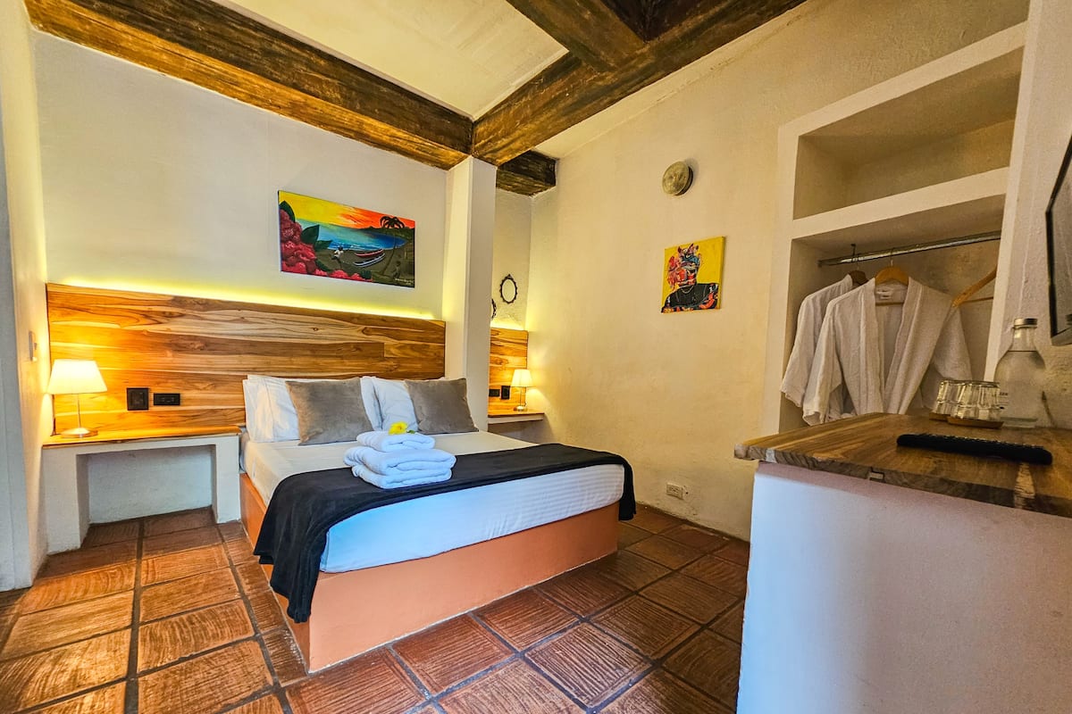 A hotel room with a wooden headboard, a bed with white linens, a small bedside table with a lamp, and a bathroom with a mirror and towel rack.