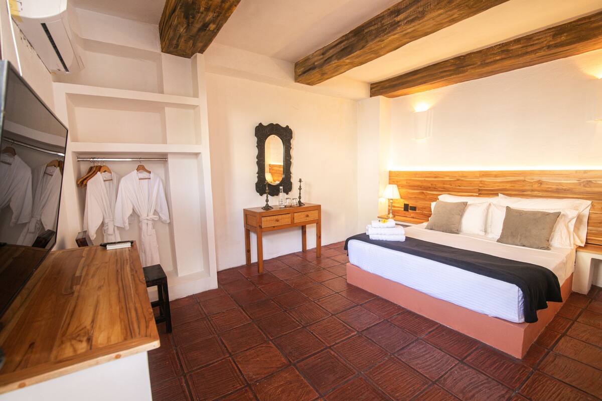 A bedroom with a wooden headboard, a bed with white linens, a wooden nightstand, a mirror, and a wardrobe with hanging robes.