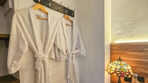 A bathroom with two white robes hanging on a rack, a wooden nightstand with a glass bottle and a lamp, and a wooden wall. - Casa Be Getsemani (Cartagena)
