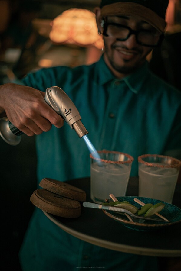 A person in a teal shirt holding a device, possibly a lighter, over a plate with a drink and a knife.