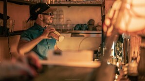 A person pouring a drink at a bar with shelves of glasses and bottles in the background. - Casa Be Getsemani (Cartagena)