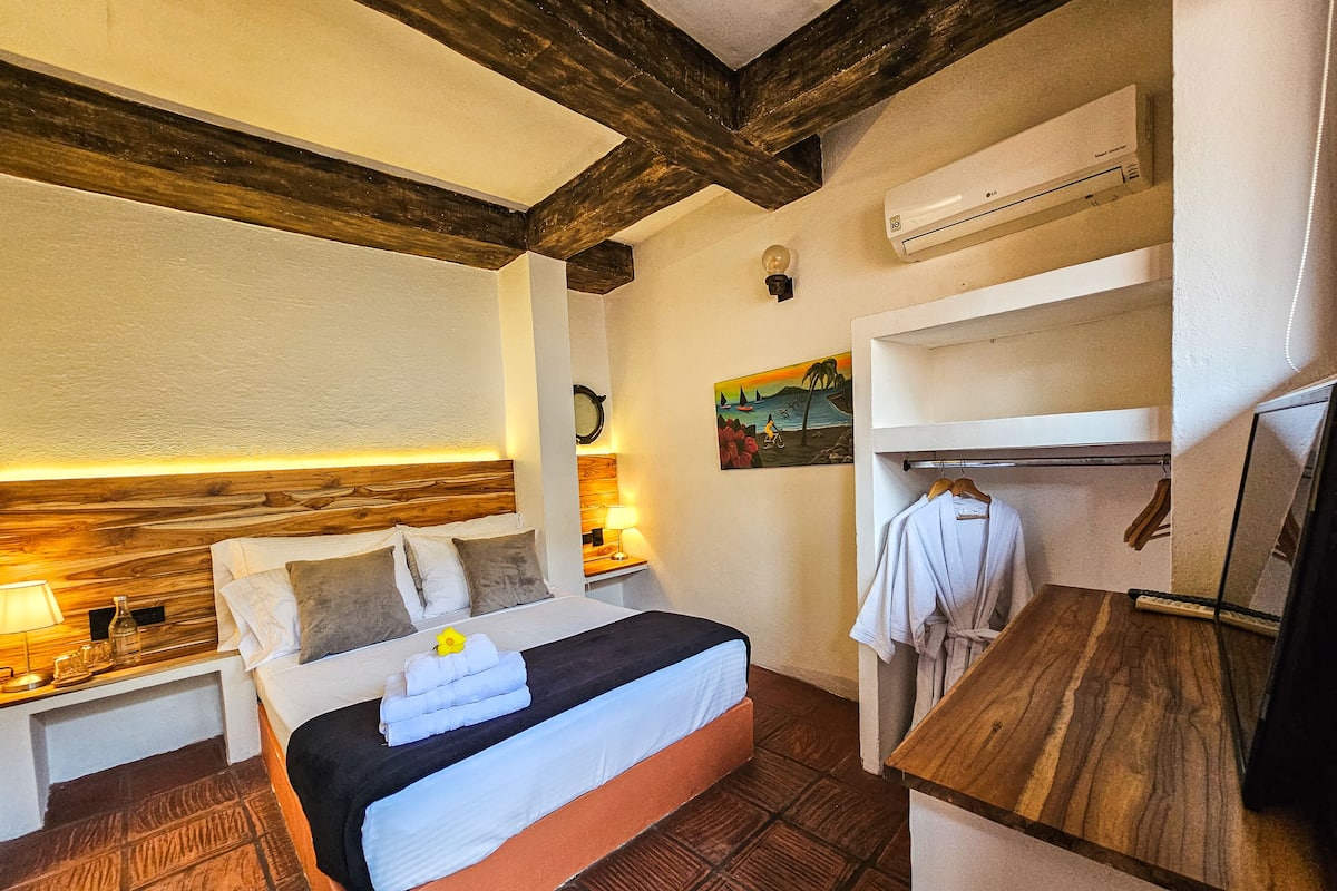 A hotel room with a wooden headboard, a bed with white linens, a nightstand with a lamp, a painting on the wall, a built-in shelf with clothes, and an air conditioning unit.