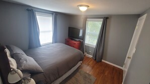 Un dormitorio con cama, televisor sobre un mueble, ventana con cortinas y puerta.