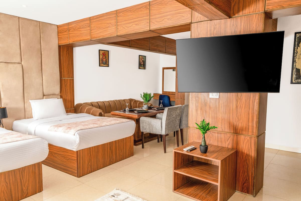 A modern hotel room with a wooden headboard, a flat-screen TV, a sofa, a dining table, and a small plant.