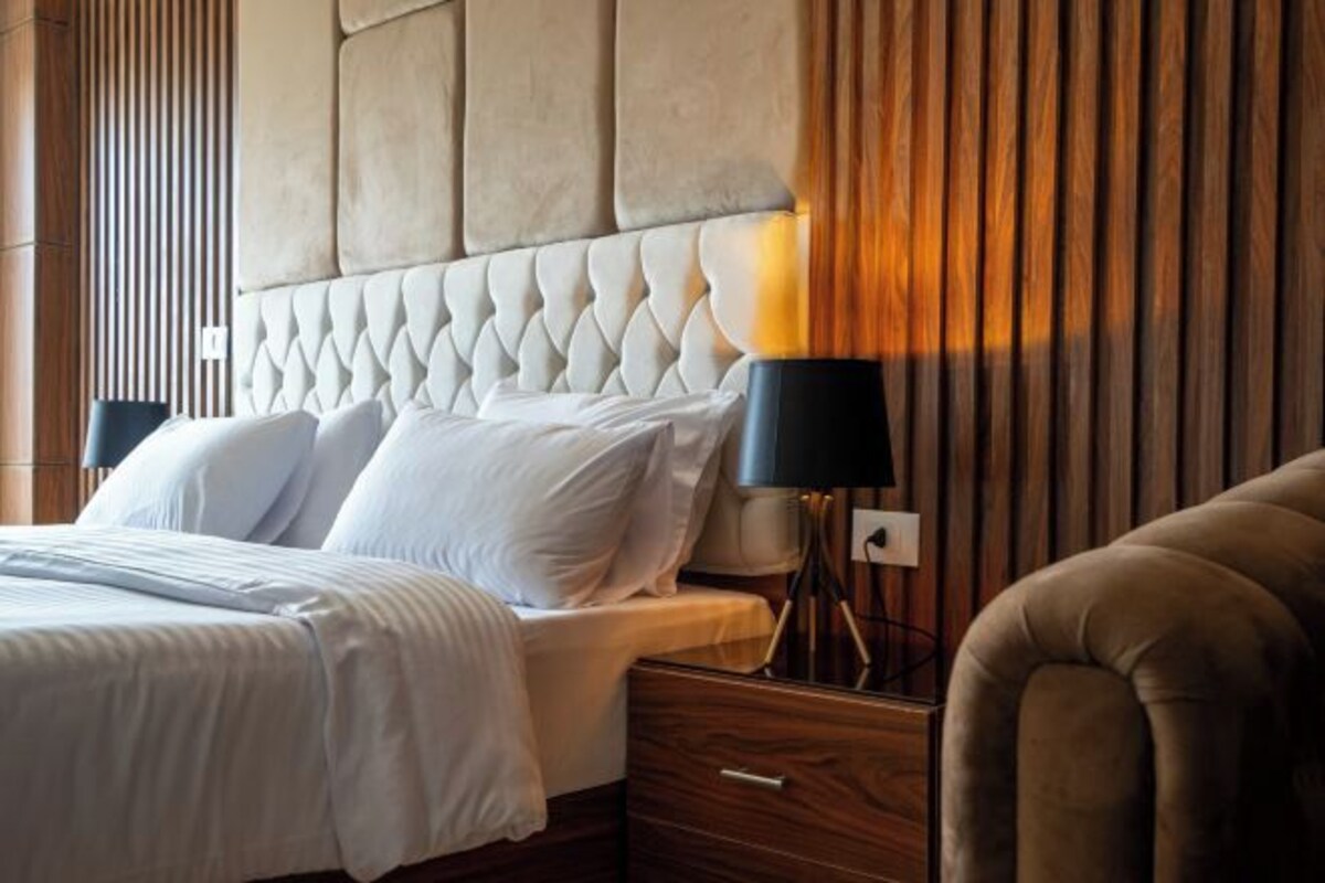 Una habitación de hotel con tablero de cabecera capitonado, una cama con ropa blanca, una mesita de noche con una lámpara y un panel de pared de madera.