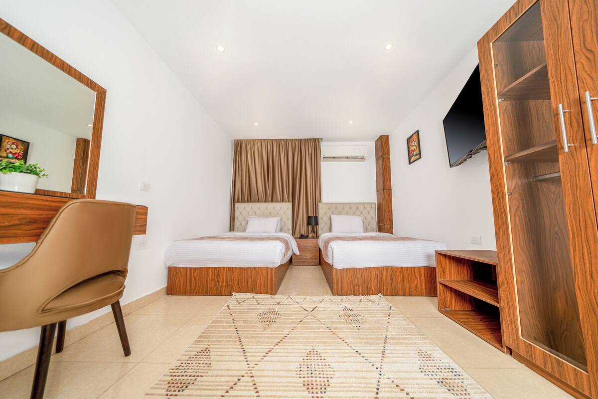 A hotel room with two beds, a wooden wardrobe, a desk, a chair, a TV, and a patterned rug.