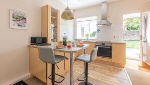 A modern kitchen with a dining area, featuring a kitchen island, bar stools, and a large window providing natural light. - Beautiful 4* Cottage in a peaceful, rural setting with Country Club membership.  (Saltash)