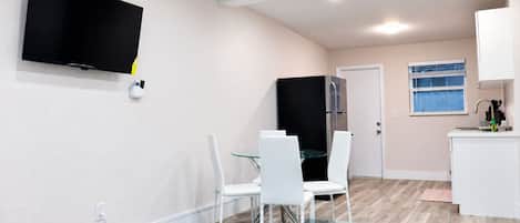 A modern kitchen with a refrigerator, a dining table with chairs, and a flat-screen TV mounted on the wall.