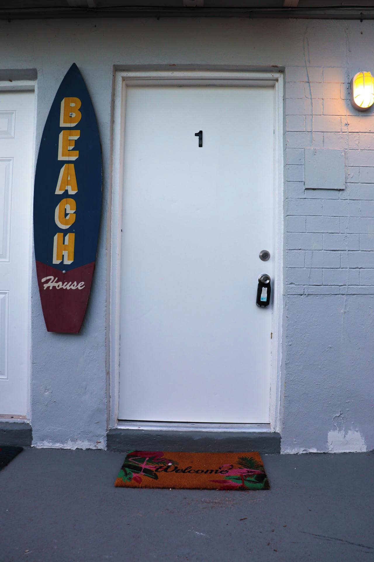 Una puerta blanca con el número 1 encima, un letrero con forma de tabla de surf que dice 'BEACH House', y un tapete de bienvenida colorido con la palabra 'Welcome'.