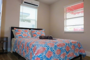 A bedroom with a bed, two windows with blinds, a bedside table, and an air conditioning unit.