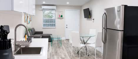 Una cocina moderna con un refrigerador de acero inoxidable, una zona de comedor con una mesa de vidrio y sillas blancas, y un sofá gris.
