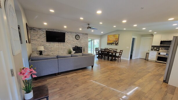 Een ruime woonkamer met een grijze bank, houten vloeren, een eettafel met stoelen, een keuken met moderne apparatuur en een flatscreen televisie bevestigd aan een bakstenen muur.
