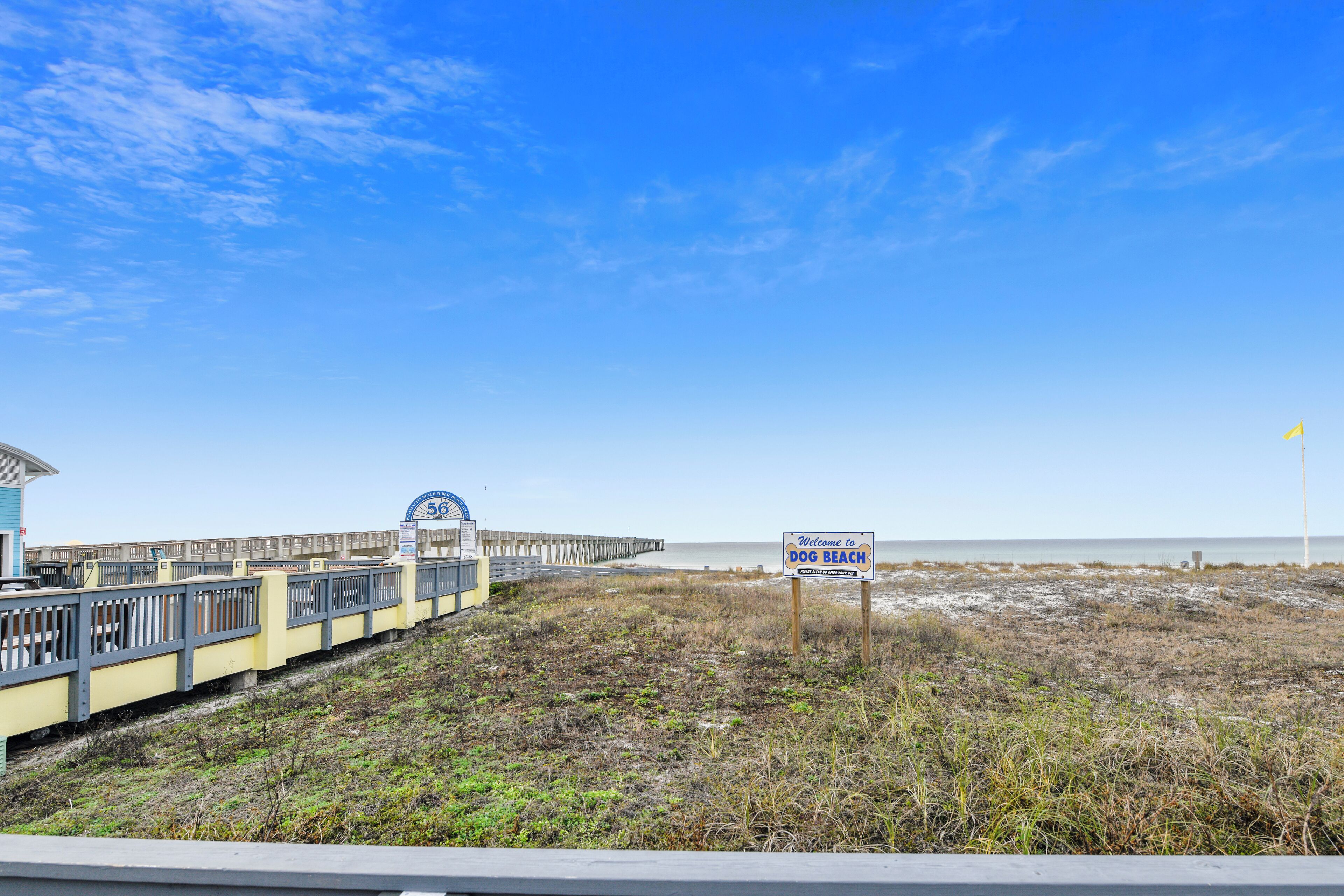 A beach area with a wooden boardwalk, a sign reading "Welcome to Dog Beach," and a pier in the background.