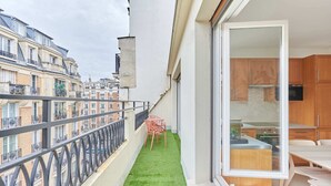 Een balkon met een kleine tafel en stoelen, uitzicht op omliggende gebouwen en een keuken met houten kastjes en een raam.