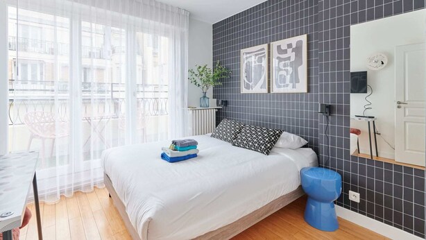 Een moderne slaapkamer met een groot bed, een nachtkastje, een blauwe kruk, een radiator en een raam met dunne gordijnen.