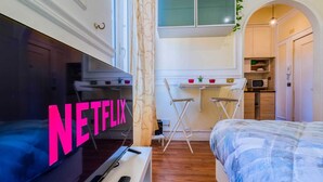 Ein kleines Zimmer mit einem Bett, einem Fernseher mit 'NETFLIX'-Logo, einem kleinen Tisch und einer Küchenzeile.
