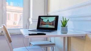 Ein Laptop auf einem weißen Schreibtisch mit Pflanze, Stuhl und Fenster mit Blick auf Gebäude.