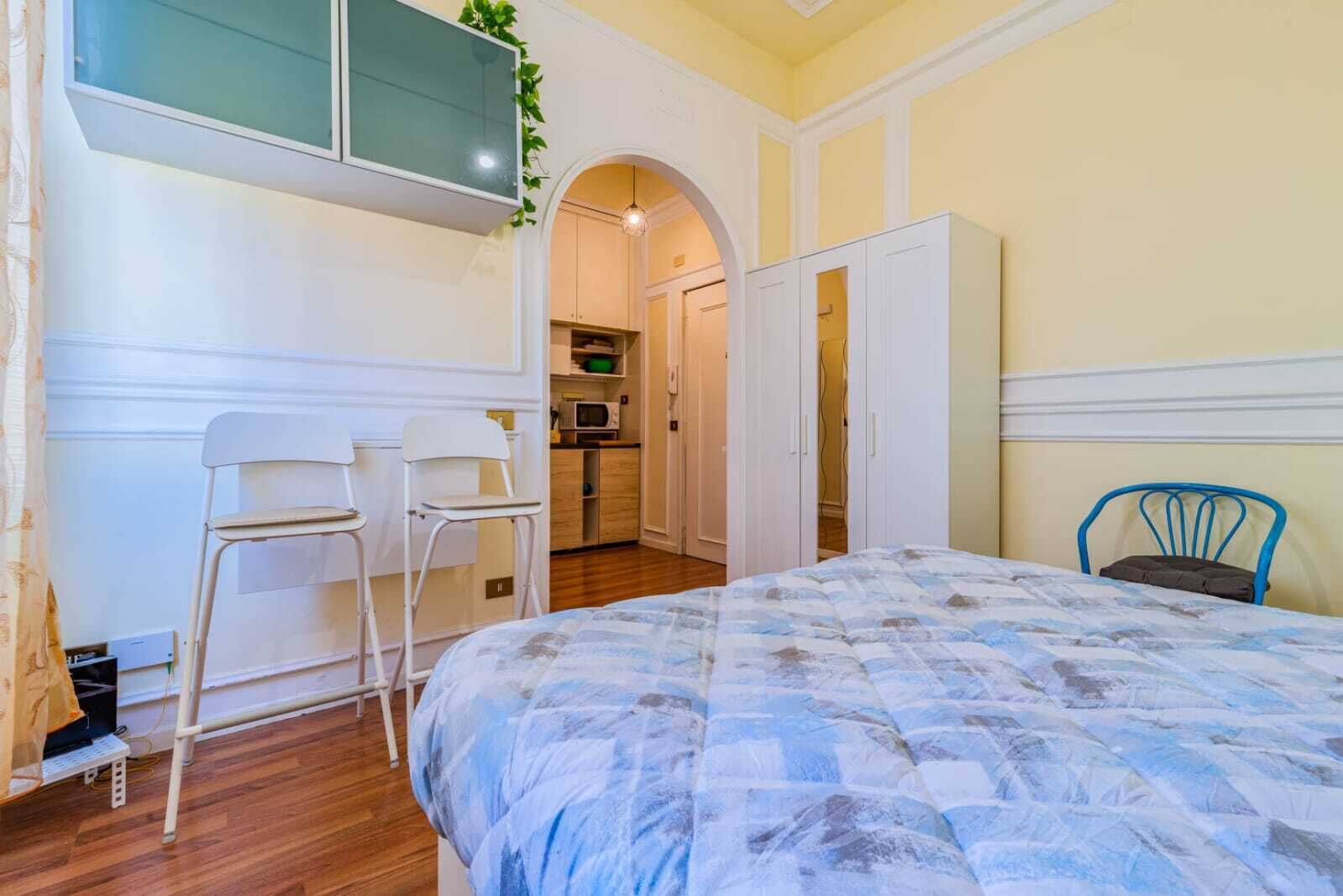 A bedroom with a bed, two chairs, a TV, and a kitchenette in the background.