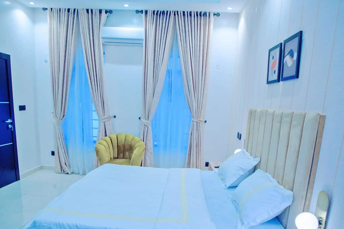 A hotel room with a bed, curtains, a chair, and a wall-mounted light.