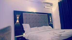 A modern bedroom with a tufted headboard, white bedding, and a wall-mounted air conditioner. - El Castillo Pretigious Hotel (Lekki)
