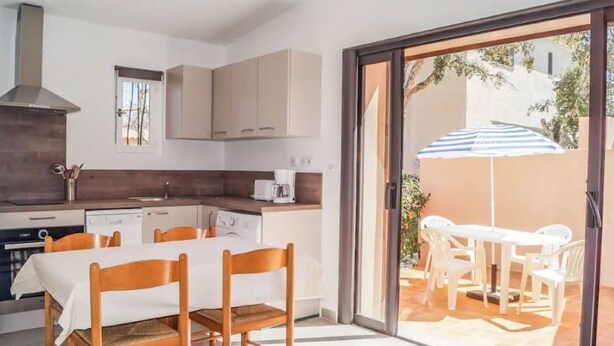 Una cocina compacta con electrodomésticos modernos, un comedor con mesa y sillas, y un piso de vidrio corredizo que da a un patio exterior con un paraguas a rayas.