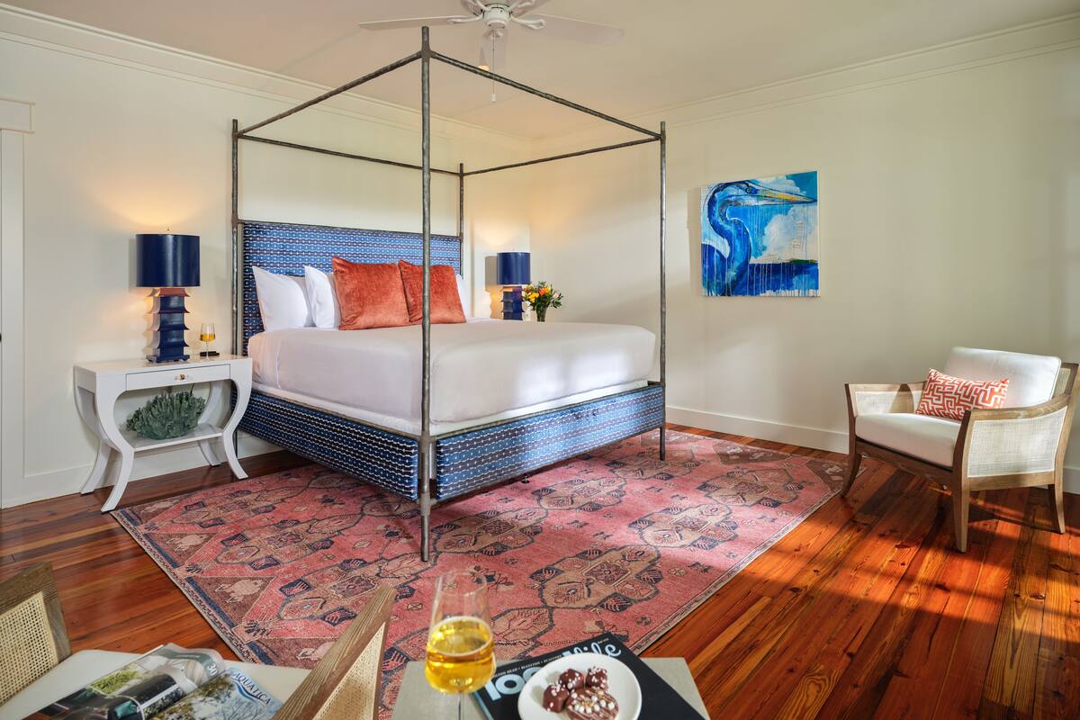 Un dormitorio con cama con dosel, una mesita de noche con lámpara, una silla, una alfombra y un cuadro en la pared.