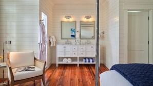 Un baño con suelo de madera, paredes blancas, un tocador blanco con dos espejos, una silla con una revista y una cama con una manta azul.