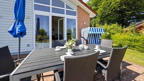 Un patio al aire libre con una mesa dispuesta para cuatro personas, rodeado de sillas de mimbre, una sombrilla azul y una silla de playa a rayas.