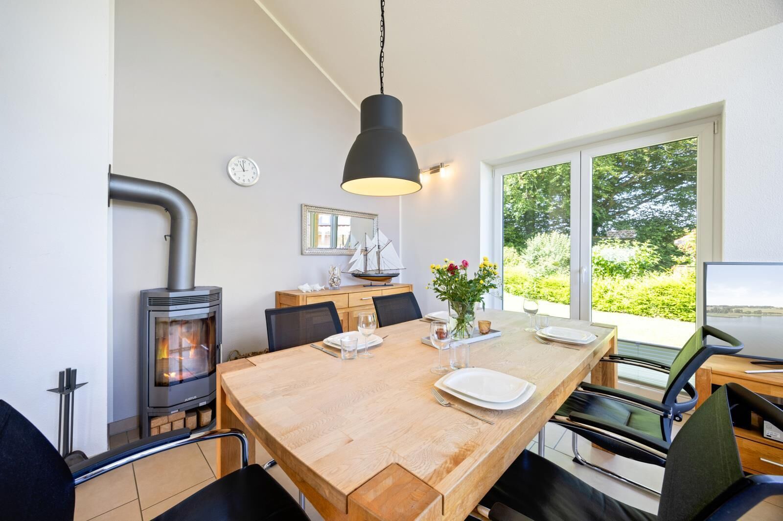 A dining area with a wooden table, chairs, a modern pendant light, a built-in wood stove, a window with a view of greenery, and a small ship model on the wall.