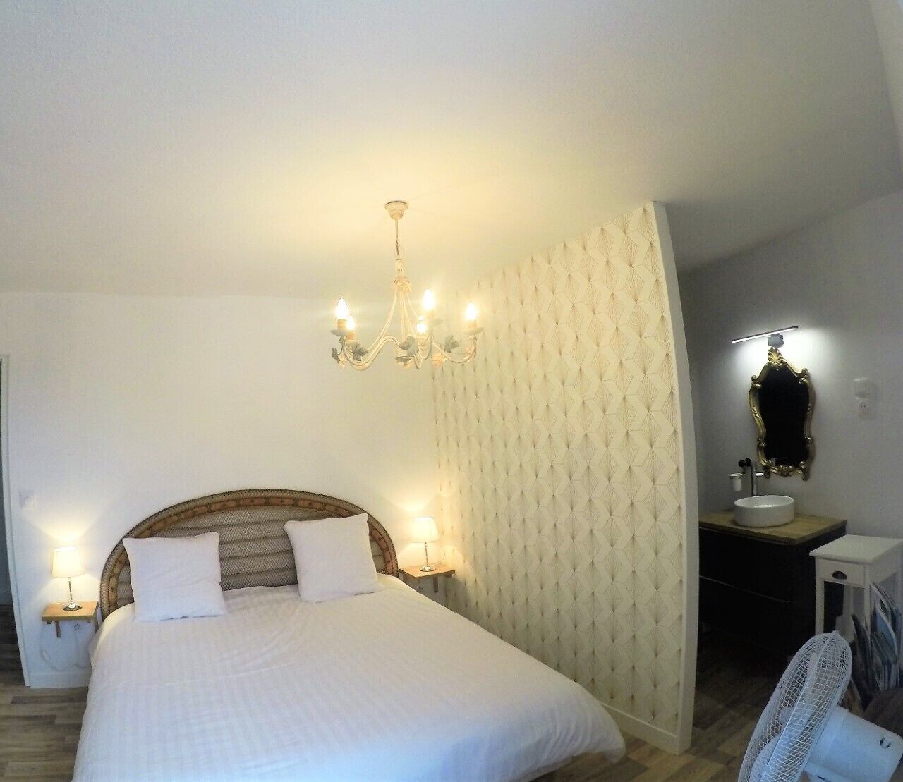 A bedroom with a large bed, a chandelier, a bedside table with a lamp, a mirror, and a fan.