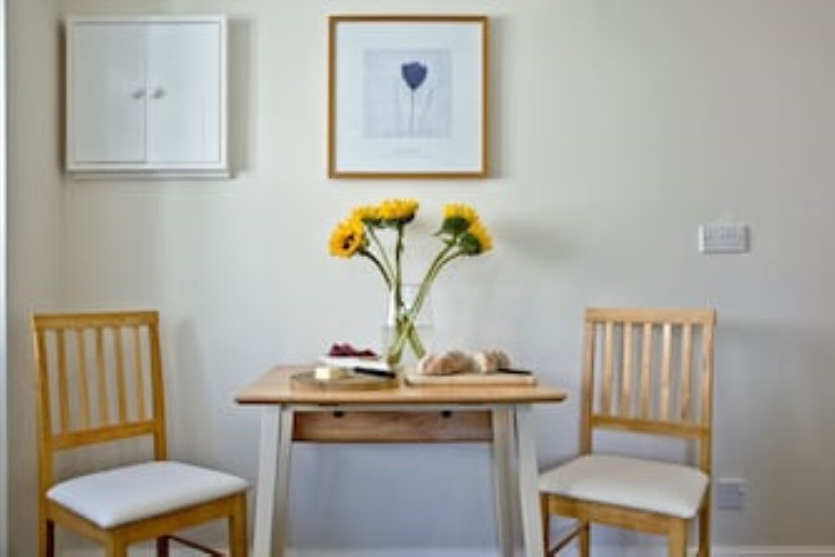 Ein Zimmer mit Holztisch, zwei Stühlen, einem gerahmten Bild an der Wand und einer Blumenvase.
