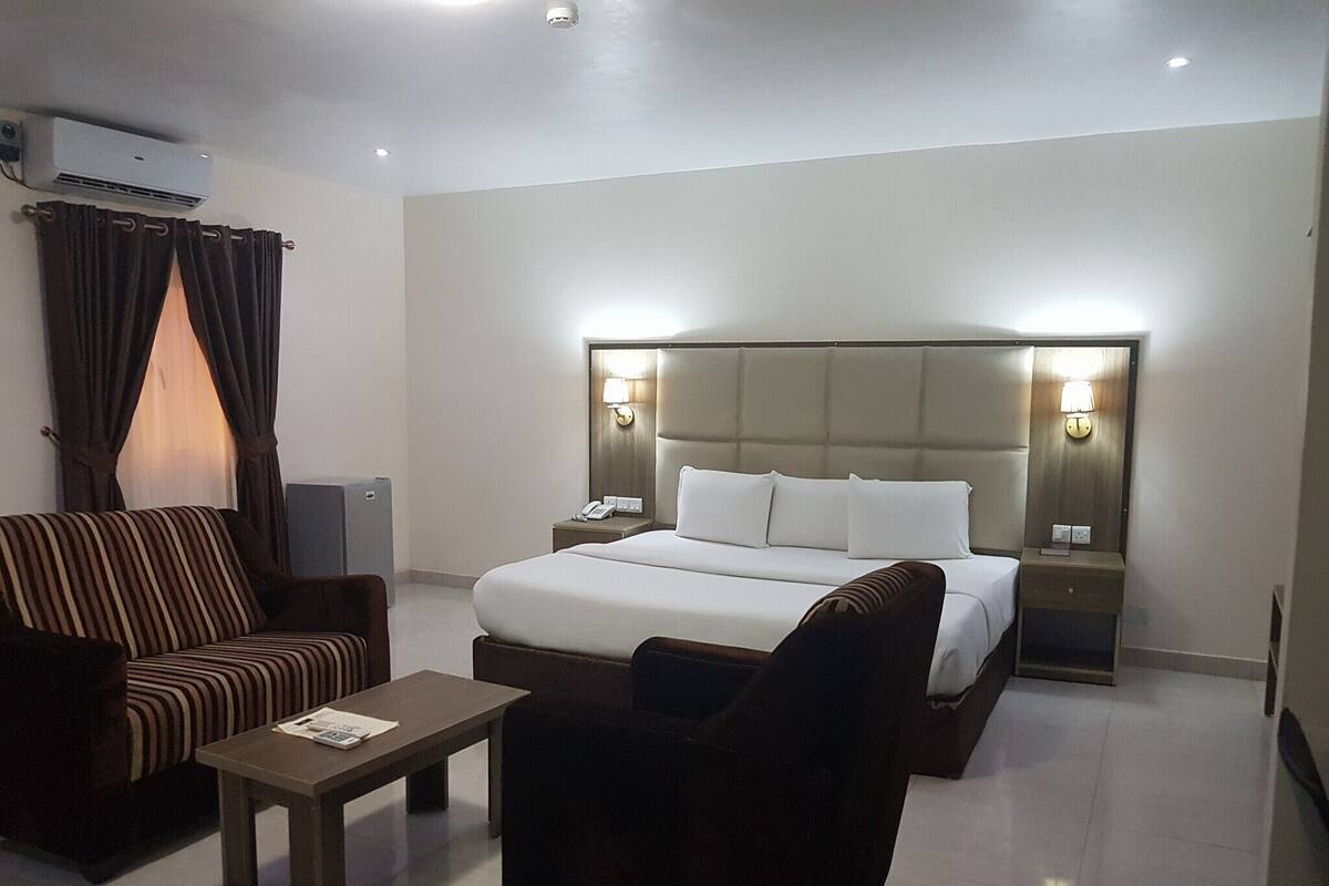 A hotel room with a large bed, two bedside tables with lamps, a sofa, a coffee table, and an air conditioning unit.