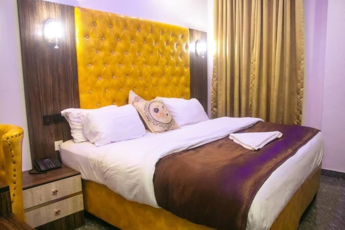 A hotel room with a tufted headboard, a bed with white linens, a brown throw blanket, a bedside table with a phone, and a yellow chair.