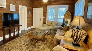 A cozy living room with wooden walls and ceiling, a large flat-screen TV, a beige sofa, a coffee table, and a patterned rug.