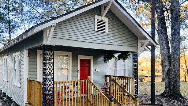 A small, single-story house with a front porch, a red door, and a staircase leading to the entrance.