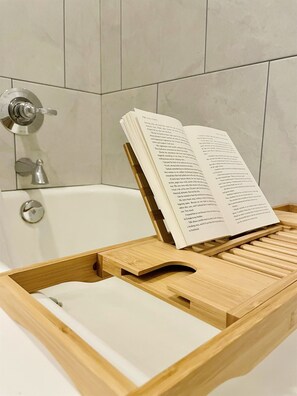 A wooden bath tray with a book open on it, placed in a bathroom with a white bathtub and a modern faucet.