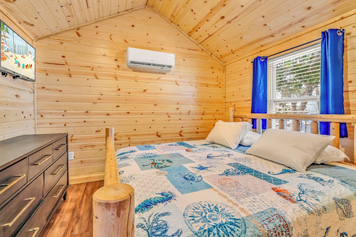 A wooden cabin bedroom with a bed, a dresser, an air conditioning unit, and a window with curtains.