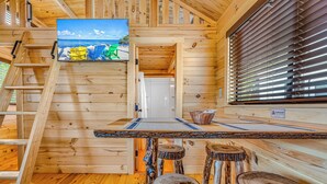 A wooden cabin interior with a wooden staircase, a dining area with a table and stools, and a window with blinds. - Cabins at Bonefish Bay (Marathon)