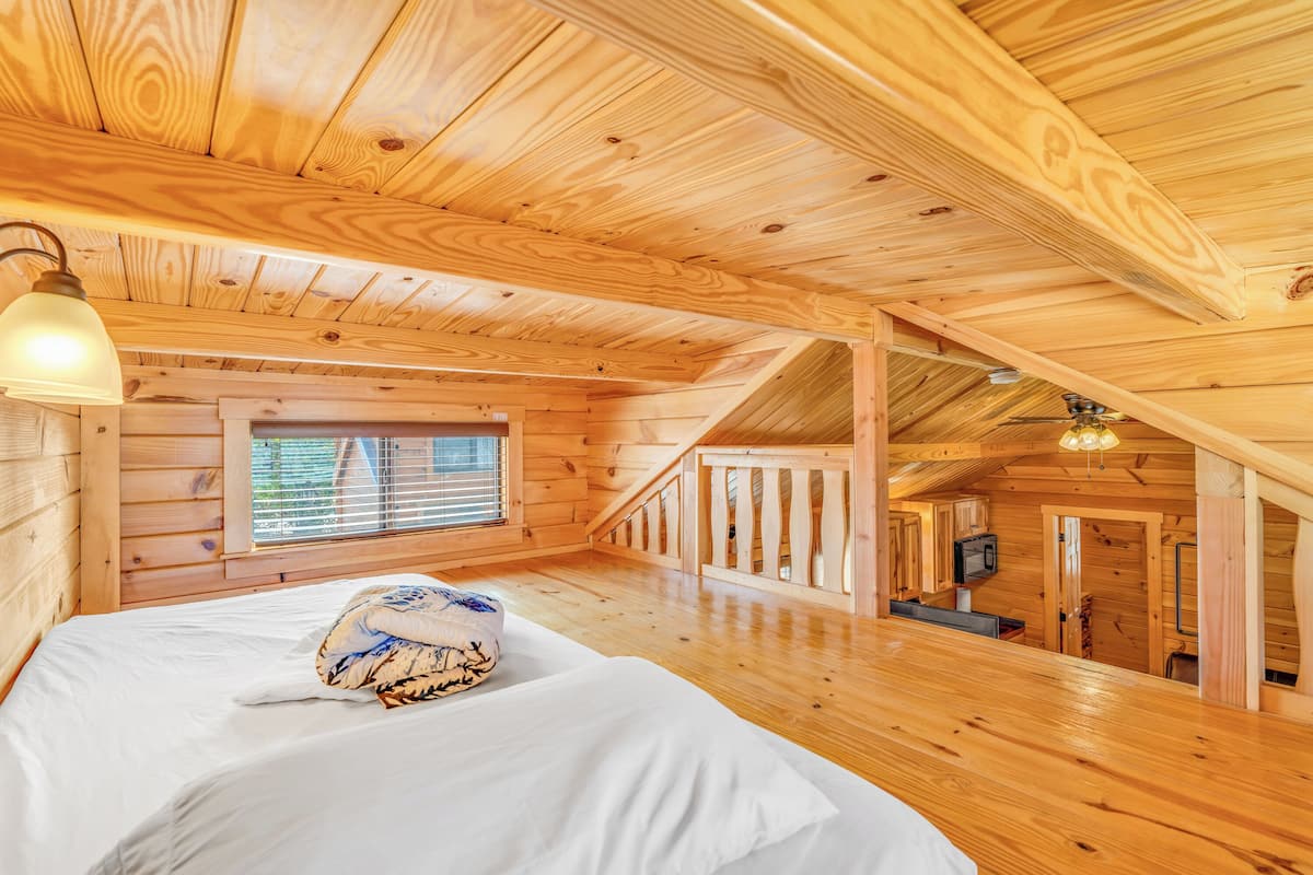 A wooden cabin interior with a bed, a window, and a staircase leading to an upper level.