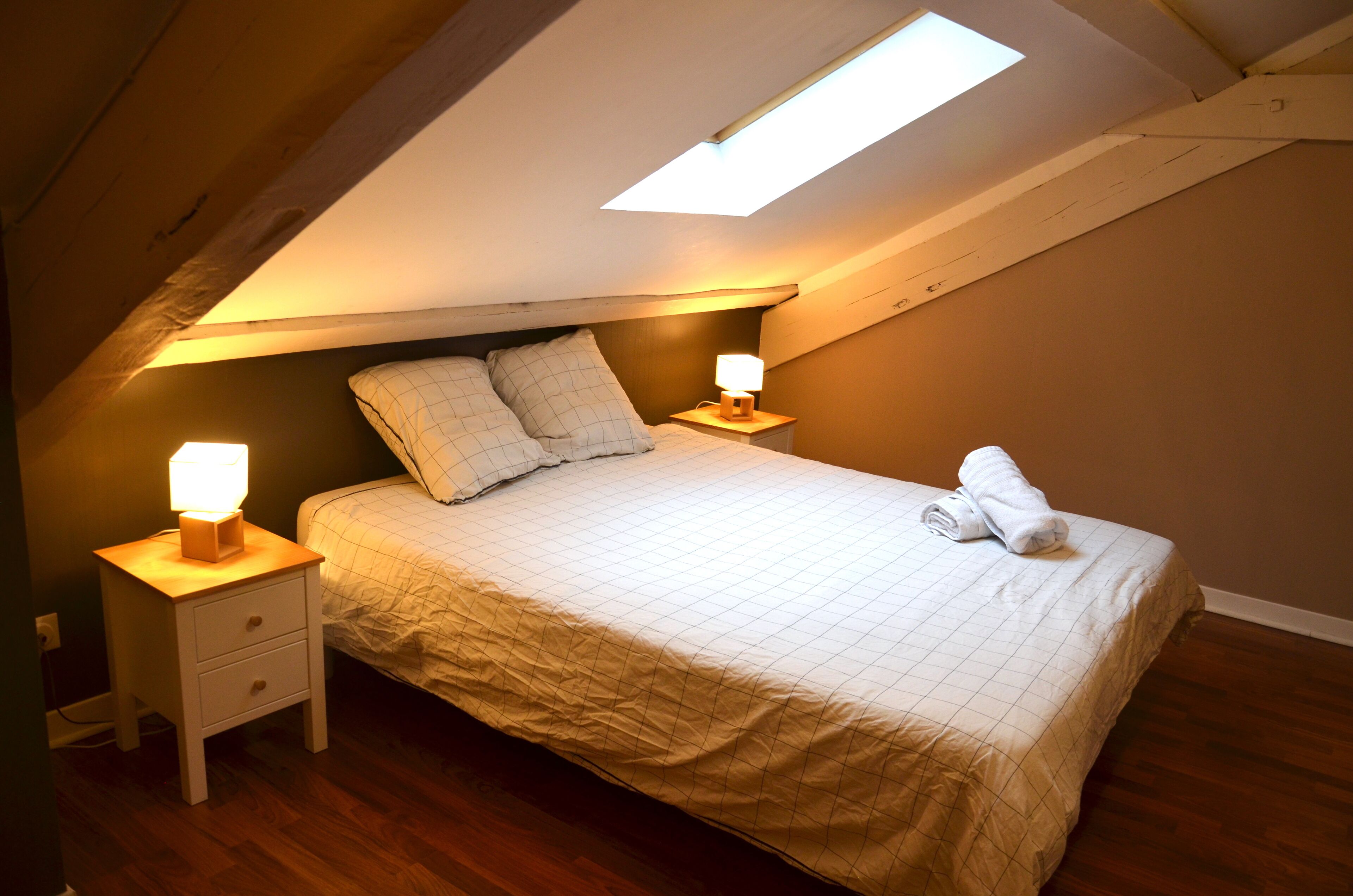 Ein Schlafzimmer mit schrägen Wänden, einem Bett mit gesteppter Bettdecke, zwei Nachttischlampen und einem zusammengerollten Handtuch auf dem Bett.