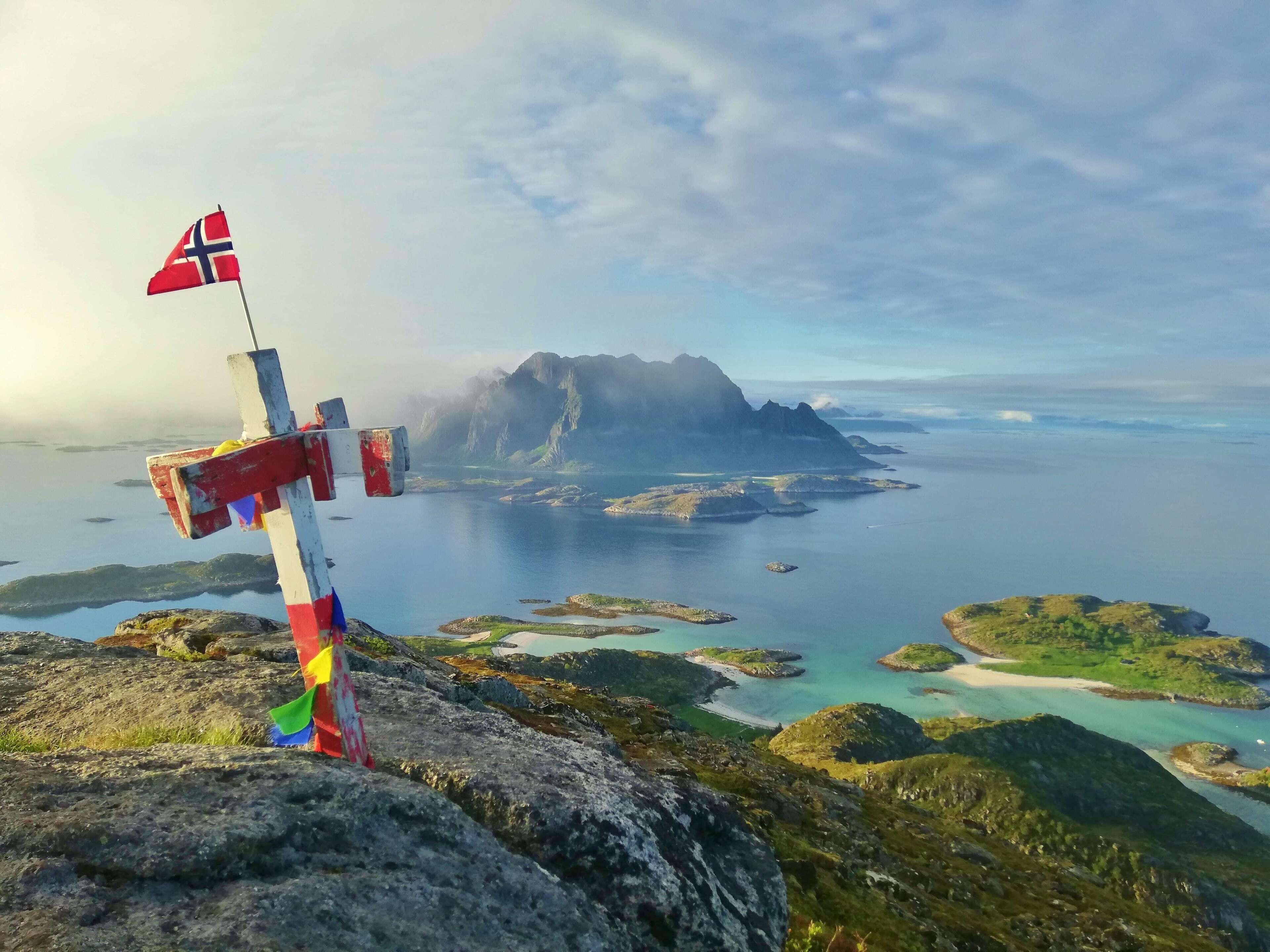 A flagpost with a Norwegian flag on a rocky outcrop overlooking a fjord with islands and mountains in the distance.