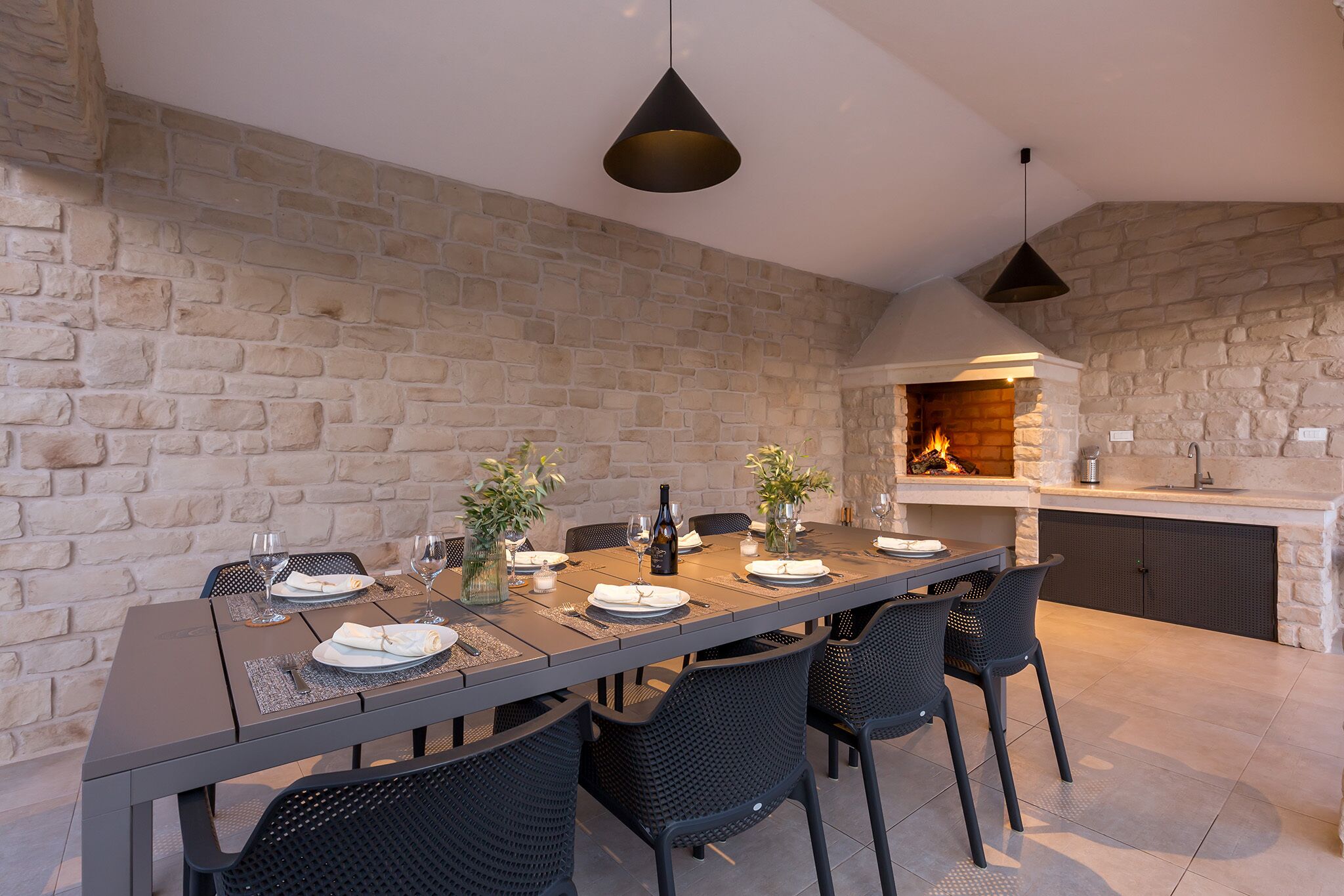A modern outdoor dining area with a stone wall backdrop, a long table set for four, and a built-in grill.