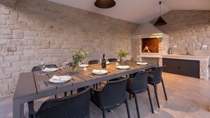 Un espace de restauration extérieur moderne, agrémenté d’un mur en pierre en arrière-plan, d’une longue table dressée pour quatre personnes et d’un barbecue encastré.