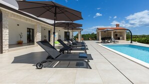 Un espace aménagé au bord de la piscine, avec des chaises longues et des parasols, une piscine et un bâtiment en pierre surmonté d’un toit en tuiles.