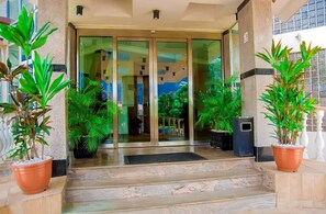 Entrada de hotel con puertas de vidrio, plantas en macetas y una escalera de mármol.